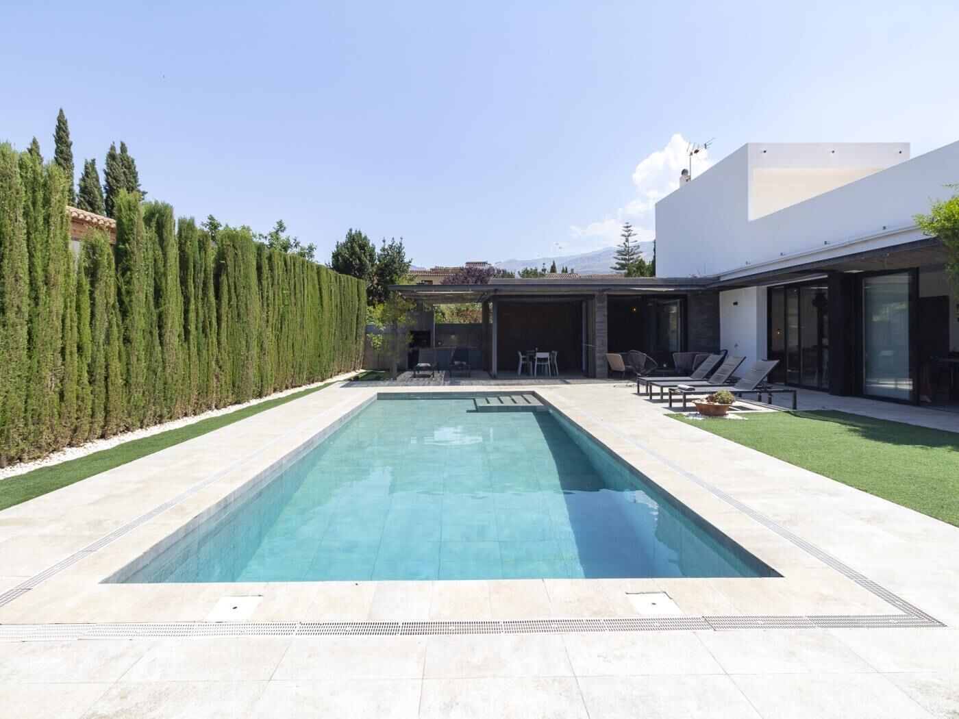 Una casa moderna con una piscina rectangular, un porche cubierto con mobiliario exterior y un césped cuidado.