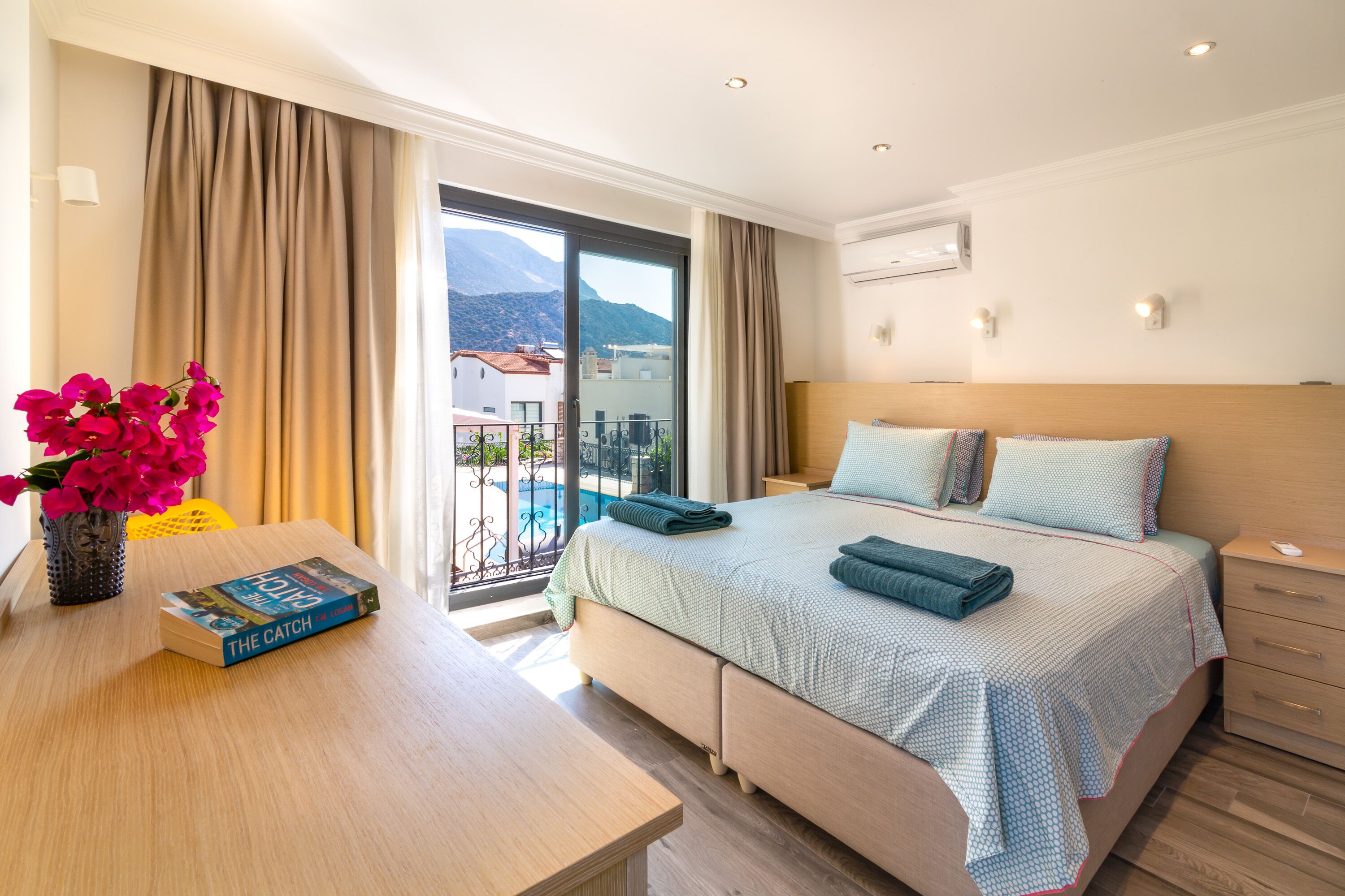 A hotel room with a bed, a bedside table with a book, a vase with flowers, and a view of a balcony and mountains.