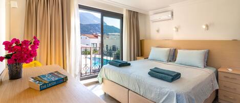 A hotel room with a bed, a bedside table with a book, a vase with flowers, and a view of a balcony and mountains.