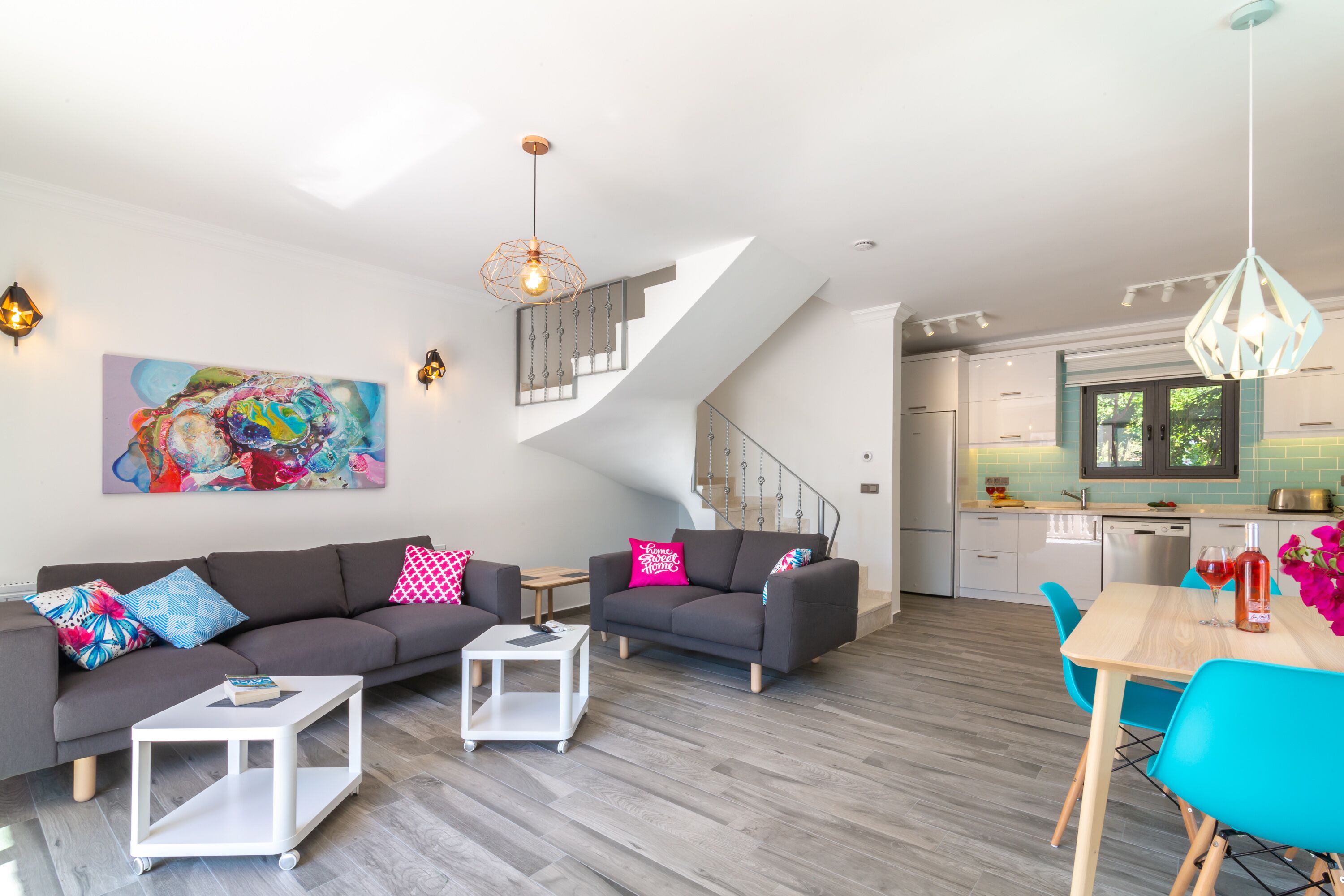 Ein modernes Wohnzimmer mit einer grauen Couch, einem weißen Couchtisch und einem Essbereich mit blauen Stühlen. Eine Treppe führt zu einer Küche mit einer grünen Spritzschutzwand.