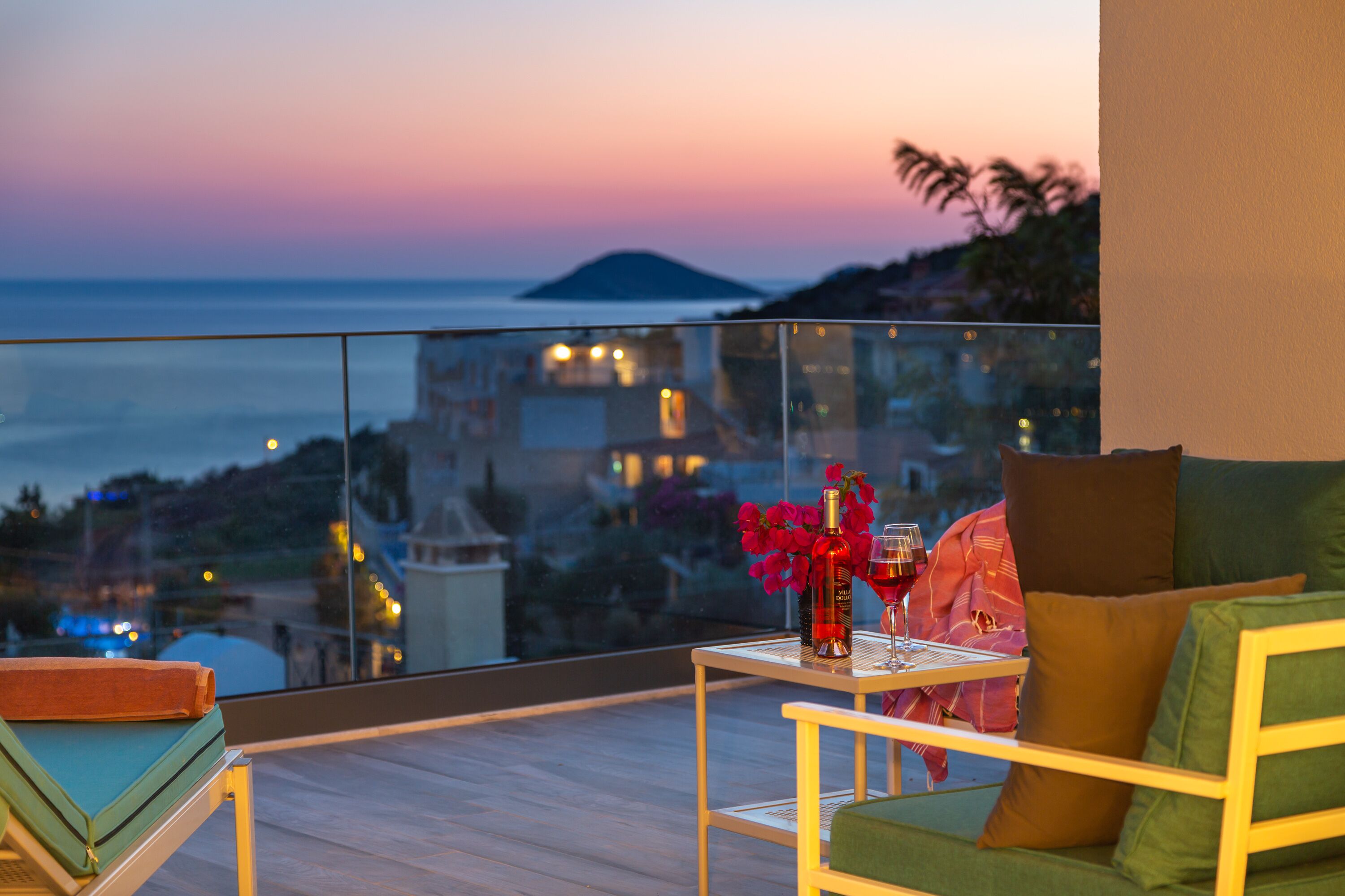 Un balcone con vista sul mare e sulla città al crepuscolo. Si vede un tavolo con una bottiglia di vino e un bicchiere.