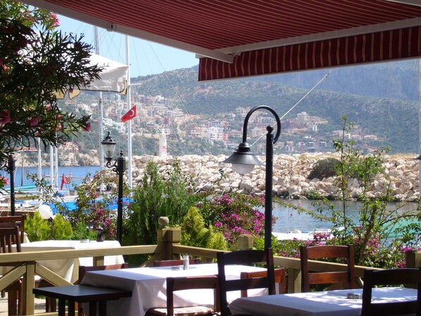 An outdoor dining area with tables and chairs, a view of a coastal town, and a flag.