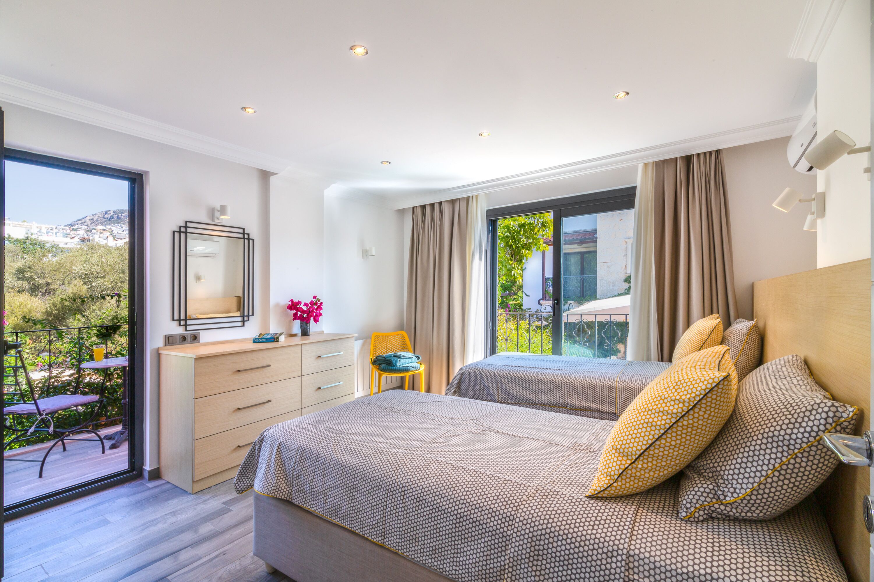 A bedroom with two beds, a dresser, a chair, a mirror, and a balcony with a view of greenery and buildings.