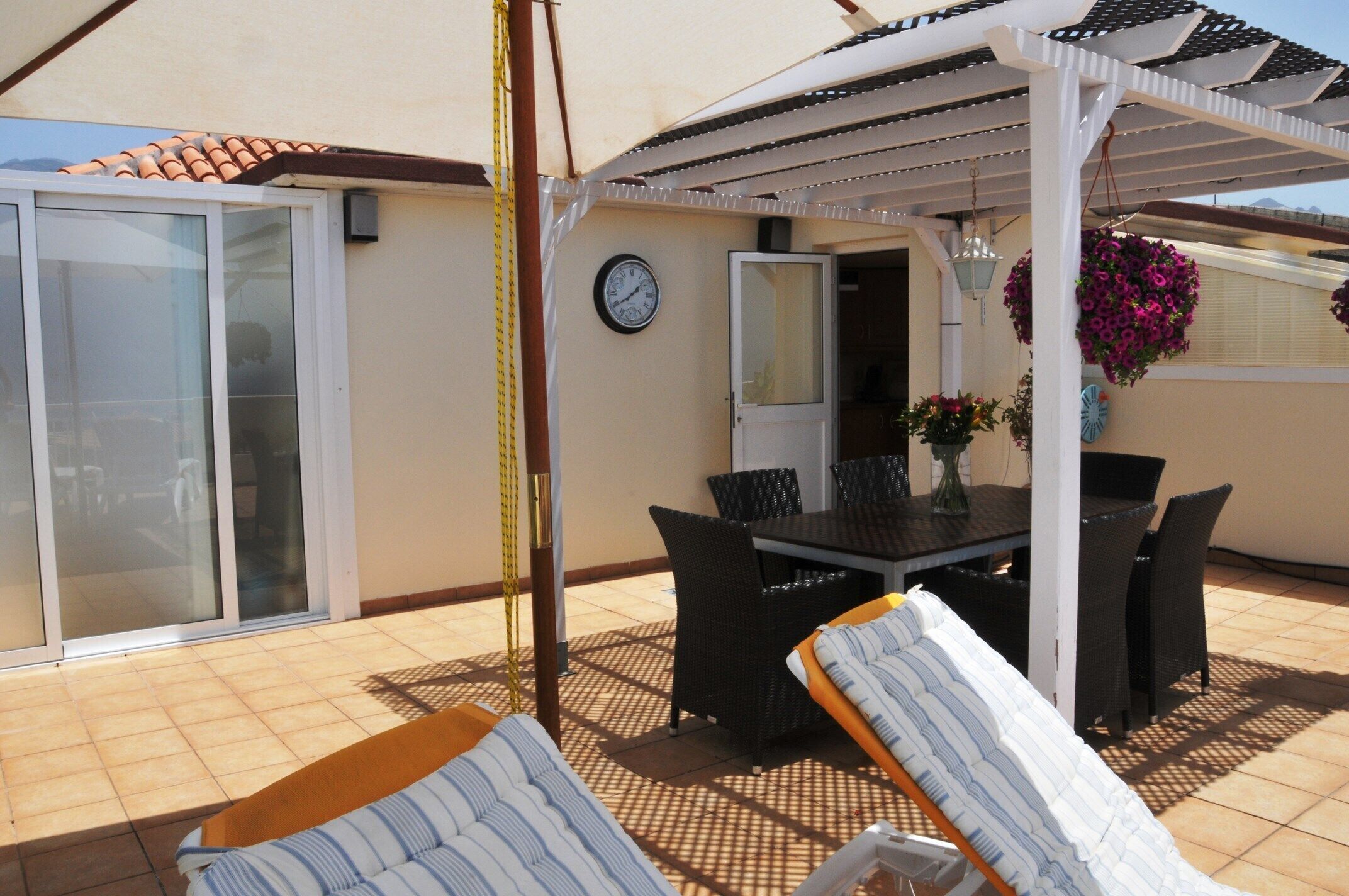 A covered patio area with a table and chairs, a clock on the wall, and potted plants.