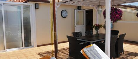 A covered patio area with a table and chairs, a clock on the wall, and potted plants.