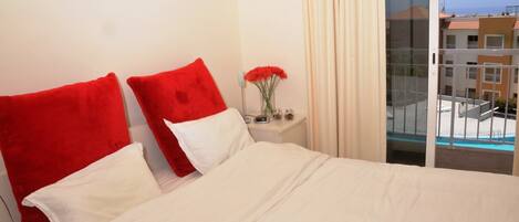 A hotel room with a bed, red pillows, a vase with flowers, a nightstand, and a view of a pool and buildings.