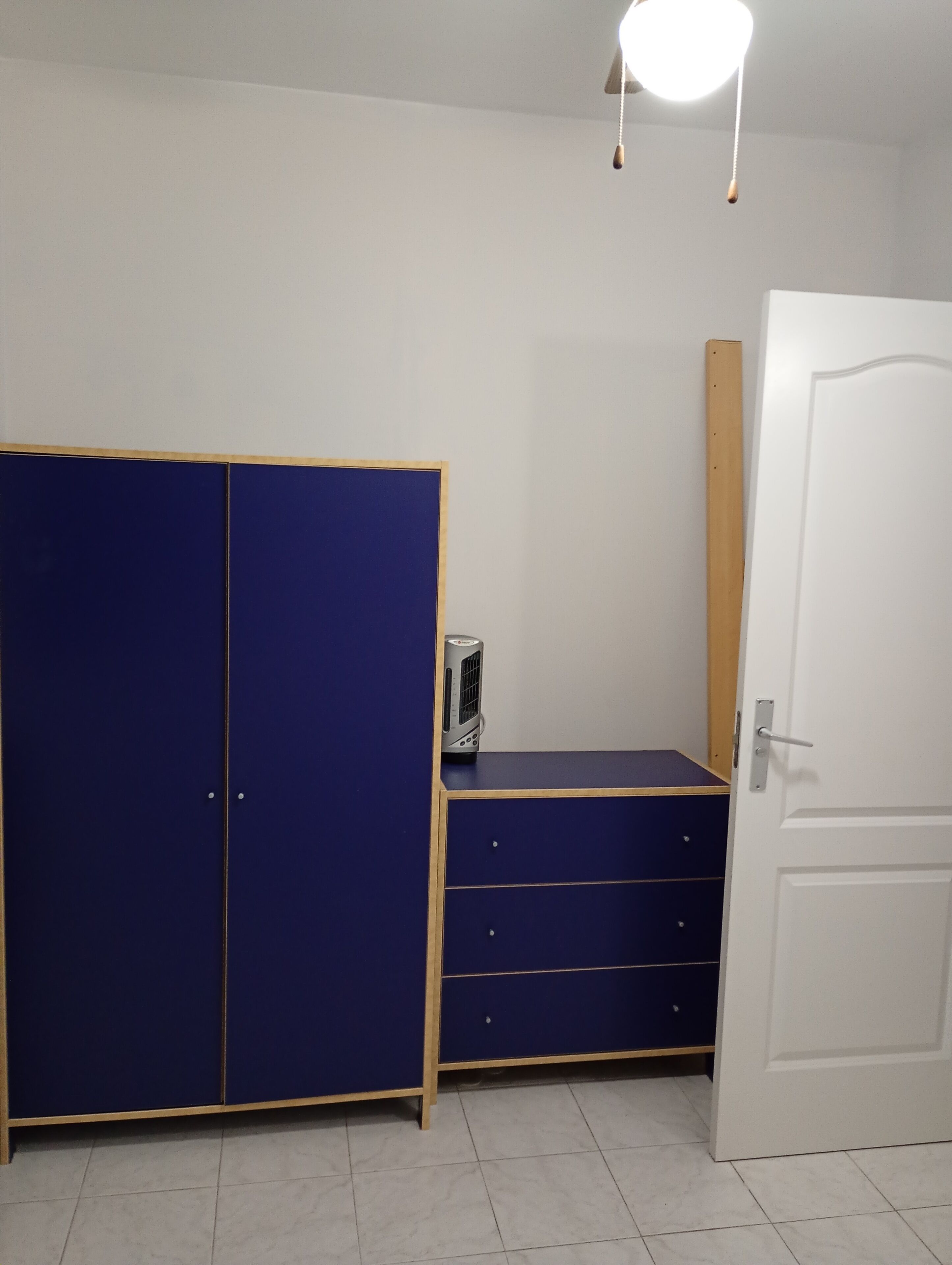 A room with a blue wardrobe, a small desk with a heater, and a white door.
