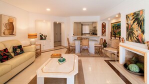 A spacious living room with a sofa, coffee table, and dining area. A kitchen is visible in the background. - Encosta do Lago, two suits Finest Apartment with Private Pool and garden. (Almancil)