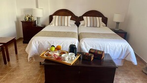 A hotel room with two beds, a wooden nightstand with a lamp, a wooden table with breakfast items, and a tray of fruits and pastries.