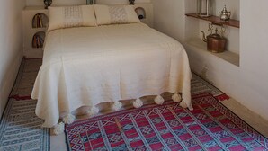 Een slaapkamer met een bed, twee bedlampen, een wandplank met decoratieve objecten, een ingelijst kunstwerk en een tapijt met een patroon.