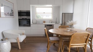 Una cocina moderna con muebles blancos, horno empotrado, microondas y refrigerador de acero inoxidable. Cuenta con una zona de comedor con una mesa redonda de madera y sillas de mimbre.