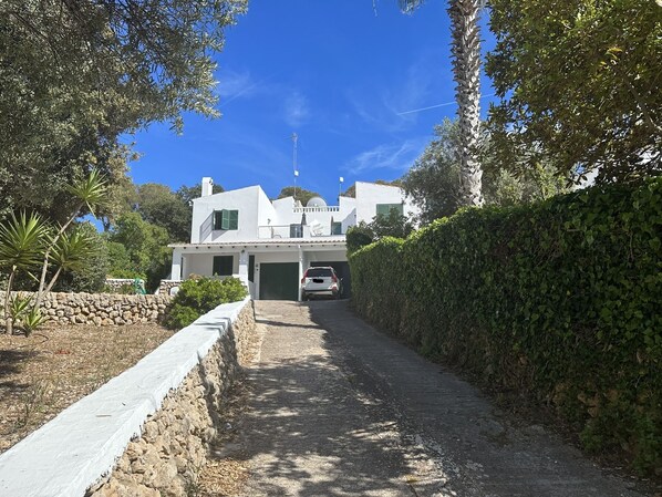 A white building with a stone wall and a paved driveway leading to a car parked in front.
