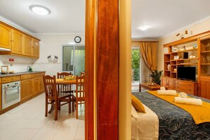 A modern kitchen with wooden cabinets, a dining area with a table set for two, a built-in oven, and a living area with a sofa and TV. - Madeira Funchal Gardens  (Funchal)