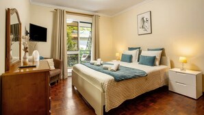A hotel room with a bed, bedside tables, a desk, a chair, a TV, a window with curtains, and a balcony view. - Madeira Funchal Gardens (Funchal)