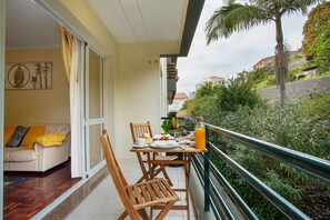 A balcony with a table set for breakfast, a sofa with cushions, and a view of greenery and buildings. - Madeira Funchal Gardens  (Funchal)
