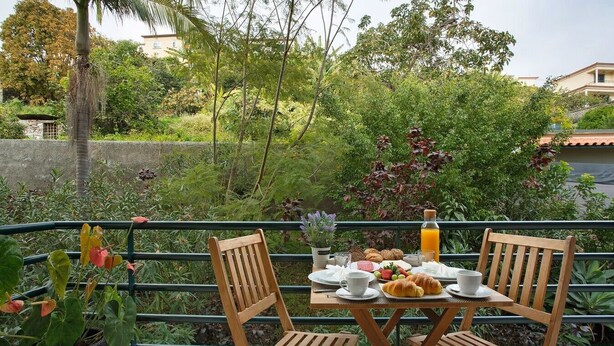 A dining table with chairs, breakfast items, and a bottle of orange juice on a balcony with a view of greenery and buildings. - Madeira Funchal Gardens (Funchal)
