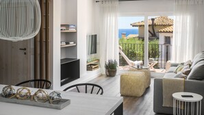 A modern living room with a sofa, a dining table, and a TV. There is a balcony with a view of the sea.