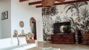 A living room with a large flat-screen TV, a white sofa, a wooden coffee table, and a wall mural of tropical foliage. - House / Villa - VILLANOVA (Villanova)