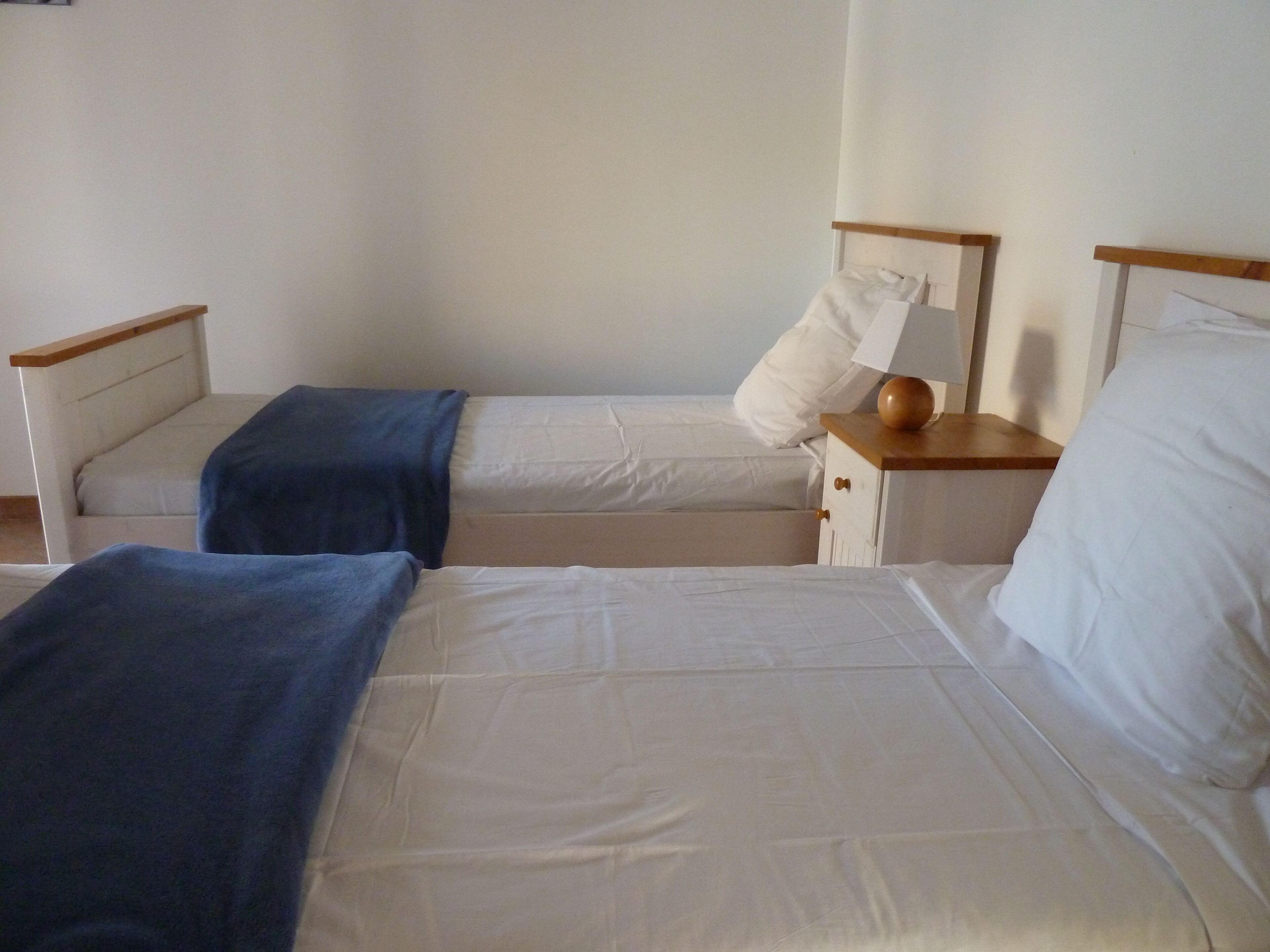 Une chambre avec deux lits, chacun doté de draps blancs et d’une tête de lit en bois. On y trouve une petite table de chevet en bois, sur laquelle sont posés une lampe et un globe décoratif.