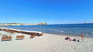 Una playa con sillas de madera y sombrillas, una orilla arenosa y una vista distante de una colina fortificada.