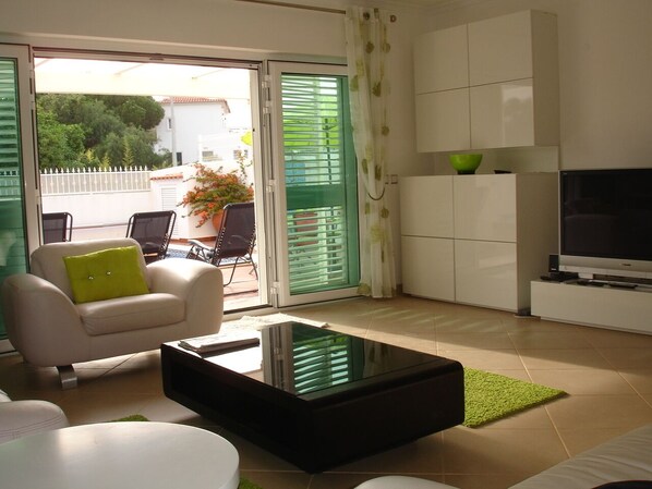 A modern living room with a sofa, a coffee table, and a TV. There is a sliding glass door leading to a balcony.