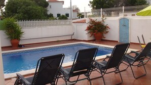 A swimming pool with three black lounge chairs, a white fence, and a tiled patio area.