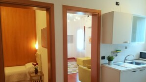 A compact hotel room with a bed, bedside table, and a small plant. A kitchenette with a sink and microwave is visible.