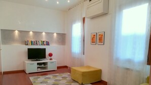 A modern living room with a flat-screen TV on a white cabinet, a yellow ottoman, a geometric rug, and two framed pictures on the wall.