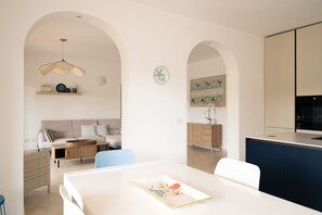 Eine moderne Küche mit weißen Schränken, ein Essbereich mit weißem Tisch und blauen Stühlen sowie ein Wohnbereich mit einer Couch und einem Couchtisch.