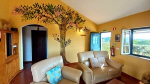 Een woonkamer met twee leren fauteuils, een houten kast, een grote muurschildering van een boom en een raam met uitzicht op groen.