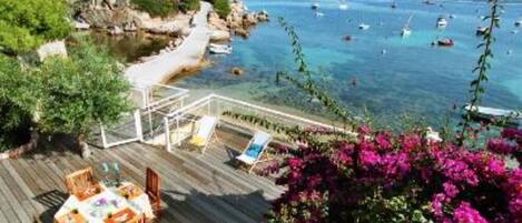 A wooden deck with a table and chairs, overlooking a coastal area with boats and greenery.
