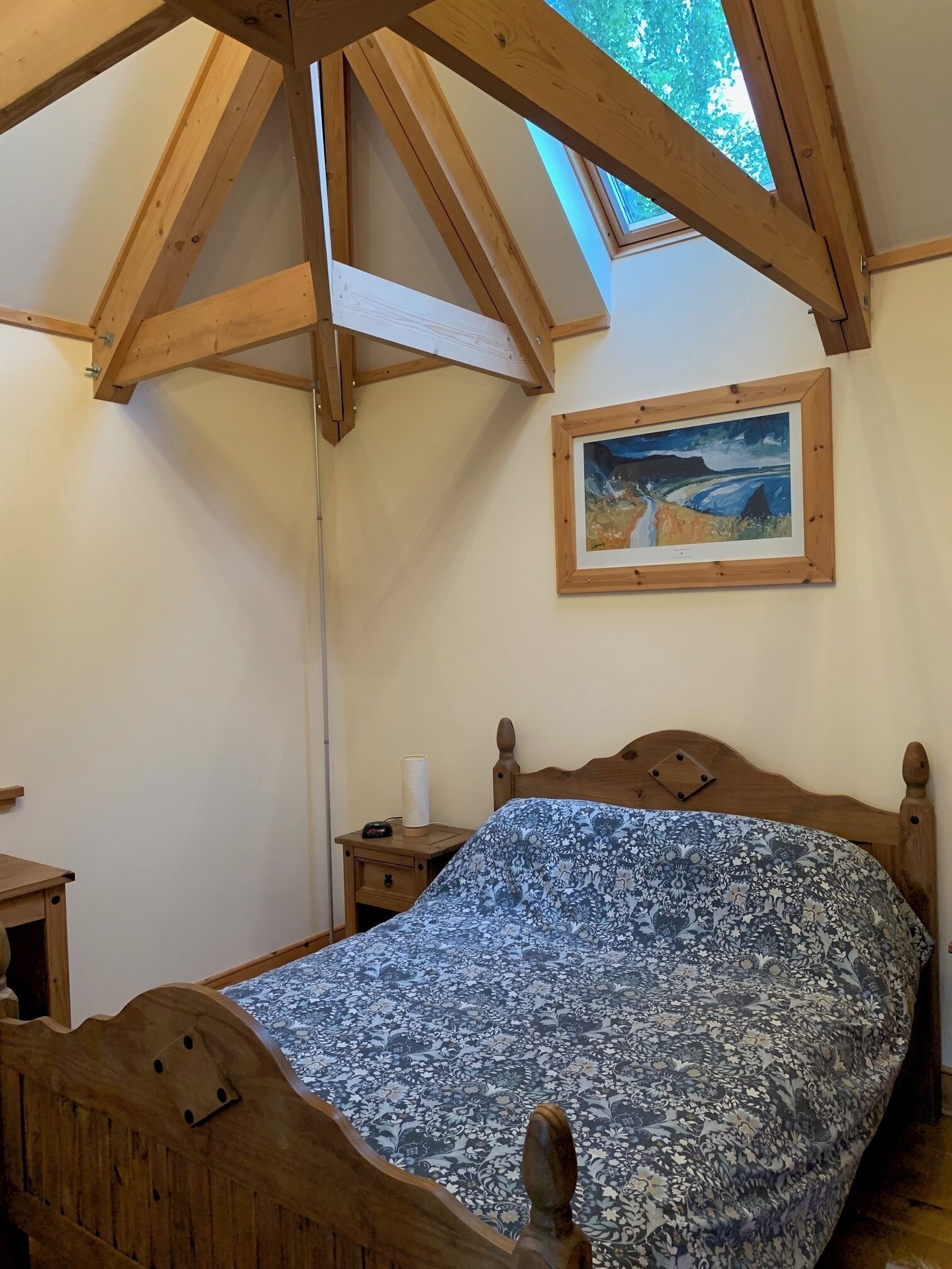 A bedroom with a wooden bed, a nightstand, a painting on the wall, and a skylight above.