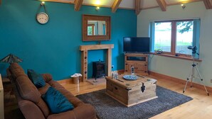 A living room with a brown sofa, a wooden coffee table, a TV on a wooden stand, a fireplace, and a large window with a view of trees. - Scottish Highlands Invergordon By NC500 Route Minimum 3 nights Any Start Day (Invergordon)