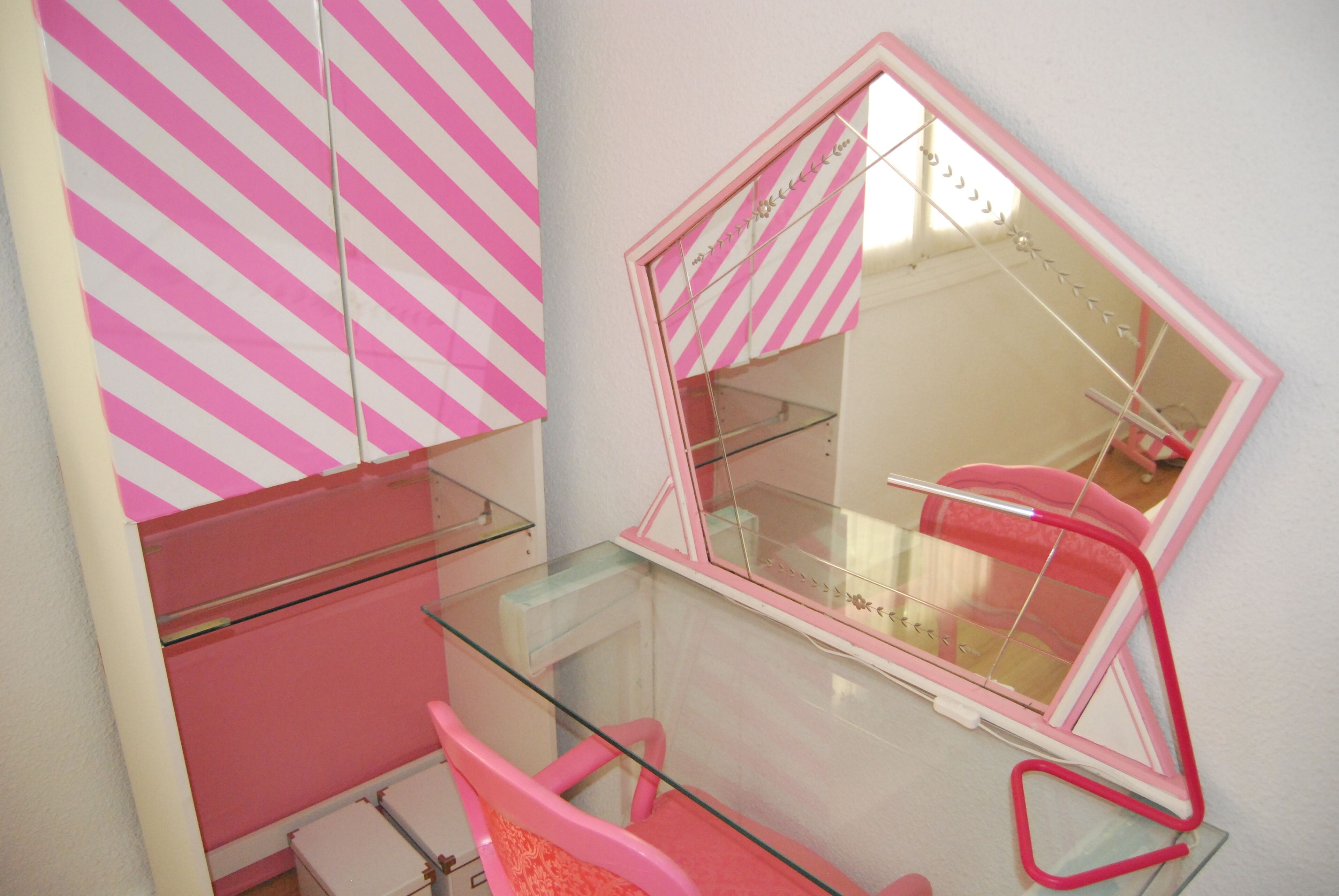 Une coiffeuse à rayures roses et blanches, dotée d’une étagère en verre et d’un miroir avec un cadre rose.
