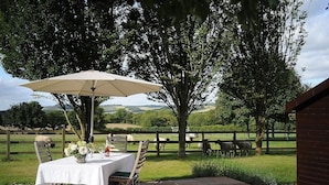 An outdoor dining area with a table set for two, a large umbrella, and a view of a fenced pasture with grazing sheep. - Romantic and peaceful cottage With Far Reaching Views  (West Meon)
