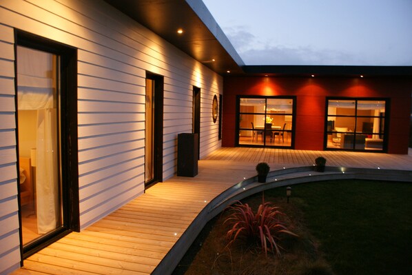 A modern house with a wooden deck, large windows, and a well-lit interior. - Single-storey house, close to the port and Kerdéniel beach. (Ploudalmézeau)