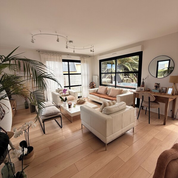 A spacious living room with a large window, a white sofa, a coffee table, and a wooden desk.