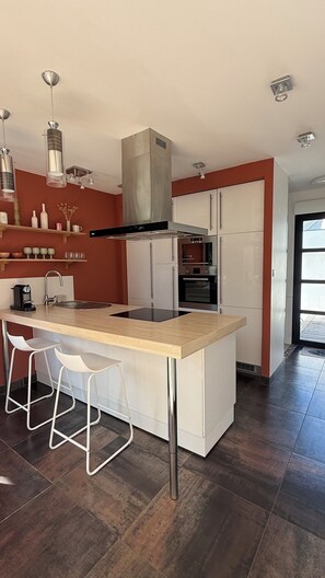A modern kitchen with a central island, stainless steel appliances, and a red accent wall. - Single-storey house, close to the port and Kerdéniel beach. (Ploudalmézeau)