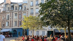 A cobblestone street with outdoor seating at cafes, surrounded by historic buildings with balconies and large windows.