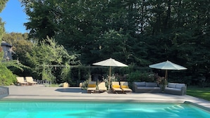 A swimming pool with lounge chairs and umbrellas, surrounded by trees and a house in the background.