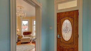 A hallway with a wooden door featuring a stained glass window, a chandelier, and a view into a living room with a sofa and a rug.