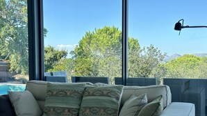Una sala de estar moderna con un sofá color beige, cojines estampados, una pequeña mesa redonda y vistas a una piscina y árboles a través de amplios ventanales.