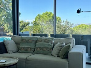 A modern living room with a beige sofa, patterned cushions, a small round table, and a view of a swimming pool and trees through large windows. - SPECTACULAR SEA VIEWS. INFINITY POOL . LIFT. (begur)