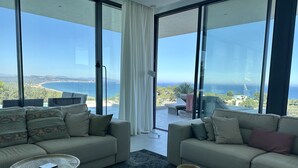 Una sala de estar moderna con dos sofás, una mesa de centro y una mesita auxiliar. Amplias ventanas ofrecen vista al mar y al paisaje distante.