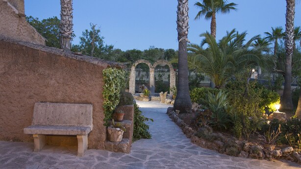 A stone pathway leading to an arched entrance, surrounded by palm trees and potted plants. - Typical Majorcan finca with pool, wireless, garden, barbecue and heating (Santanyi)