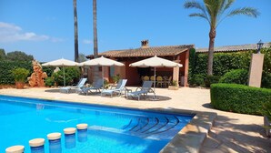 A swimming pool with lounge chairs and umbrellas, surrounded by palm trees and a building with a tiled roof. - Typical Majorcan finca with pool, wireless, garden, barbecue and heating (Santanyi)