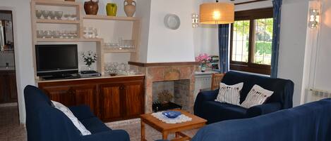 A living room with a fireplace, blue sofas, a television, and shelves with glassware.