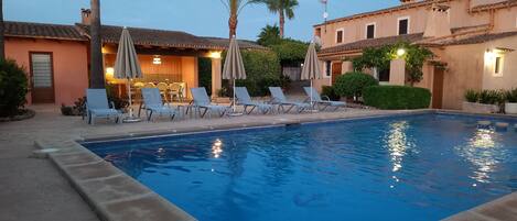 A swimming pool with lounge chairs and umbrellas, surrounded by a building with a tiled roof and a door.