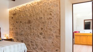 A bedroom with a stone wall, a bed with a checkered bedspread, a nightstand with a lamp, and a view into the kitchen with wooden cabinets. - Typical Majorcan finca with pool, wireless, garden, barbecue and heating (Santanyi)