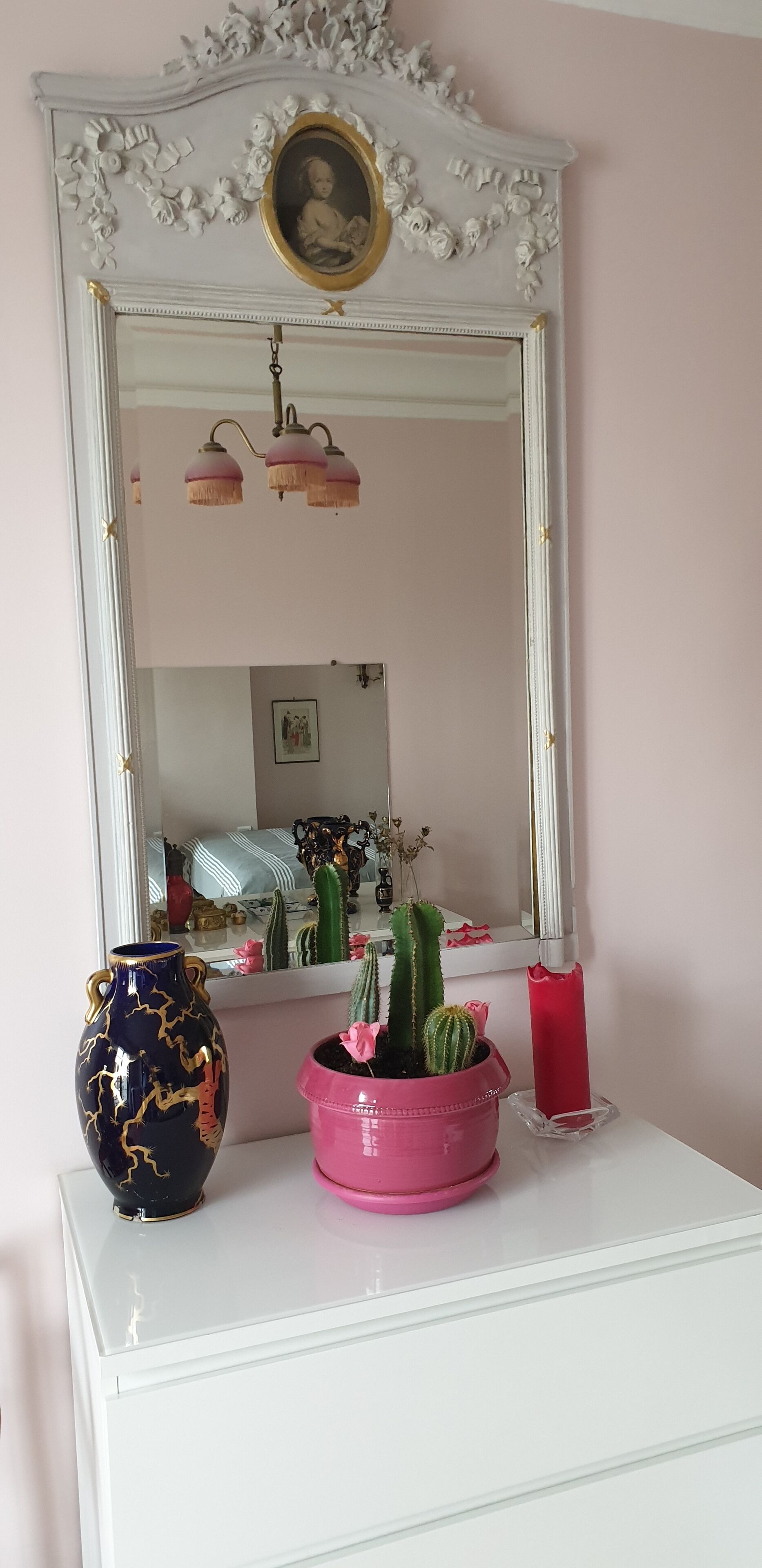 A white dresser with a mirror above it, a vase, a pink pot with a cactus, and a red candle.
